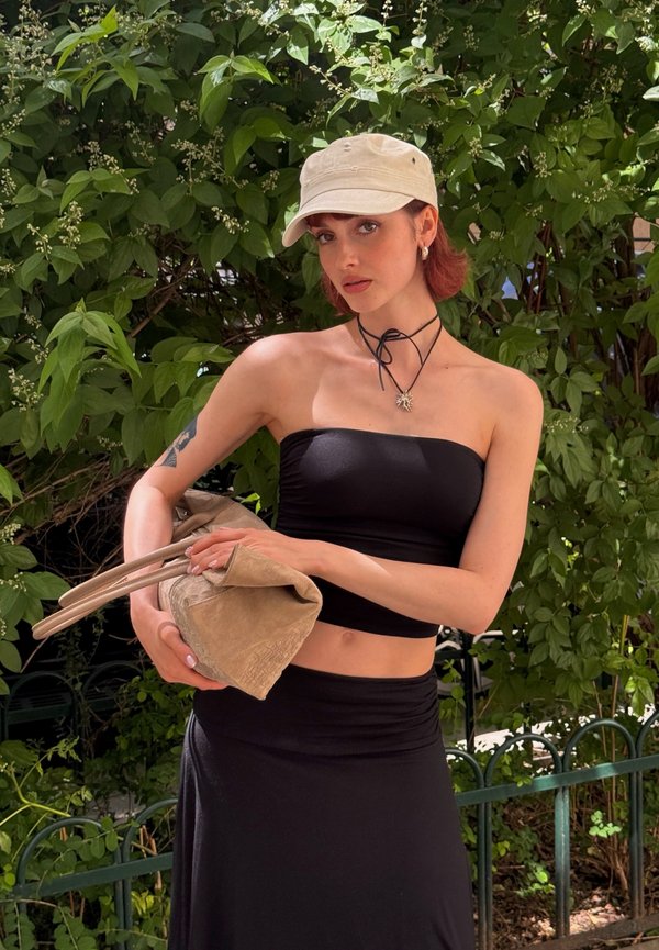 Ung kvinde i sort tube top og nederdel, der holder en beige håndtaske, iført lys kasket og solvedhængs halskæde, står udendørs nær grøn løv.