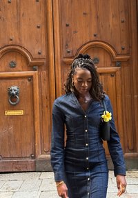 Farkkumekko pitkällä hihalla ja napeilla, tyylitelty mustalla käsilaukulla ja keltaisella kukka-aiheisella korostuksella. Taustalla puiset ovet.