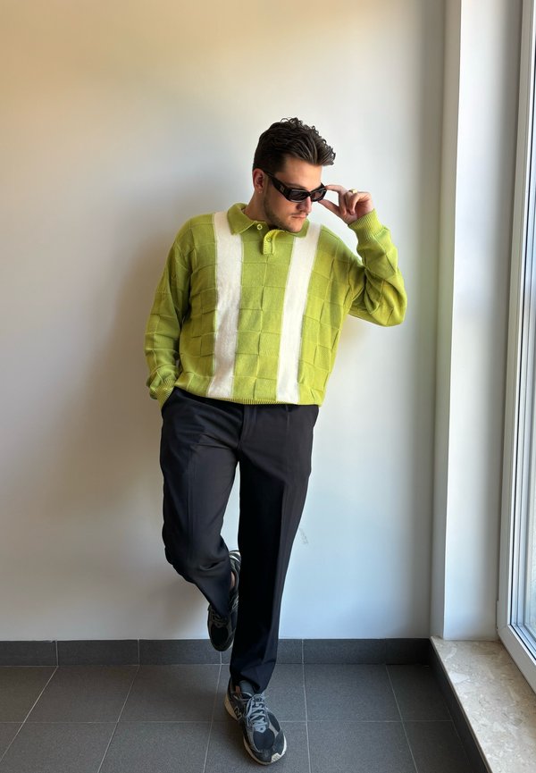 Zielony sweter z dzianiny w białe paski, luźny krój, połączony z czarnymi spodniami i czarnymi sneakersami. Model opiera się o ścianę, nosi okulary przeciwsłoneczne.