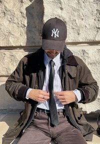 Brown outerwear with a black hat, a white pinstriped shirt, and a black tie. Hands positioned at the neck adjusting the tie. Grey stone background.