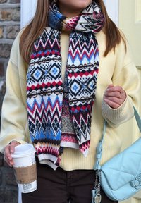 Personne portant un pull jaune et une écharpe colorée à motifs, tenant une tasse de café dans une main et un sac matelassé bleu clair dans l'autre.