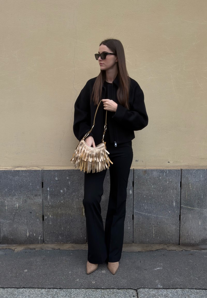 Femme en tenue noire et lunettes de soleil tenant un sac à main à franges dorées, debout contre un mur beige avec une base en pierre grise.