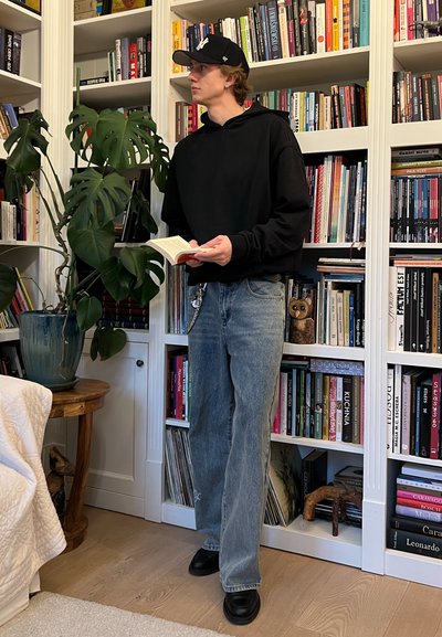 Joven con una sudadera negra y jeans azules holgados, sosteniendo un libro, se encuentra al lado de una planta grande y frondosa y estanterías llenas de libros.