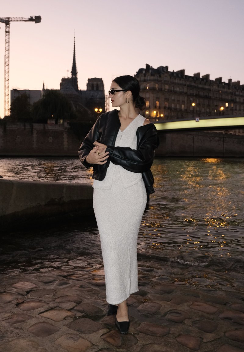 Witte getextureerde jurk met een zwart leren cropped jack, gedragen met zwarte schoenen, tegen de achtergrond van een rivier en historische gebouwen bij schemerlicht.