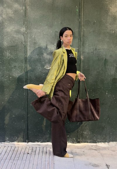 Modelo vistiendo una chaqueta amarilla texturada, un top corto negro, pantalones sueltos marrones y zapatos amarillos pálidos, sosteniendo un bolso de mano oscuro.