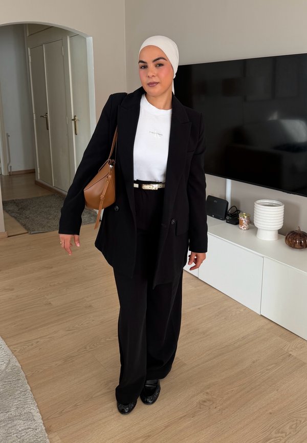 Woman wearing a white headscarf, black blazer, white shirt, black trousers, and black shoes standing in a living room with a TV and white cabinet.
