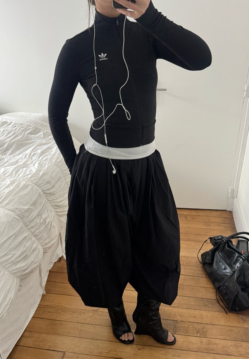 Persoană purtând un top negru Adidas cu fermoar, fustă largă neagră, cizme negre cu degetele descoperite și căști albe, realizând un selfie în oglindă în interior.