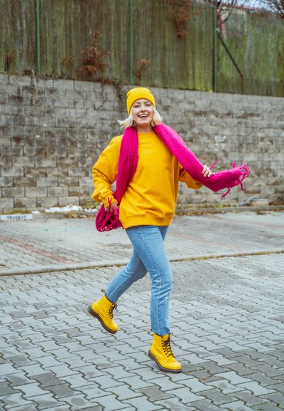 Sudadera amarilla, gorro a juego y bufanda rosa. Jeans azules y botas amarillas con cordones. Entorno al aire libre con una superficie pavimentada texturizada.