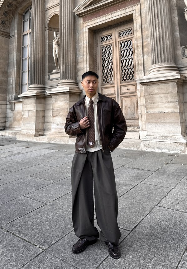 Ruda odinė striukė dėvima per šviesią marškinių palaidinę ir rudą kaklaraištį, derinama su plačiomis pilkomis kelnėmis ir tamsiais batais; stovintis prie akmeninės sienos.