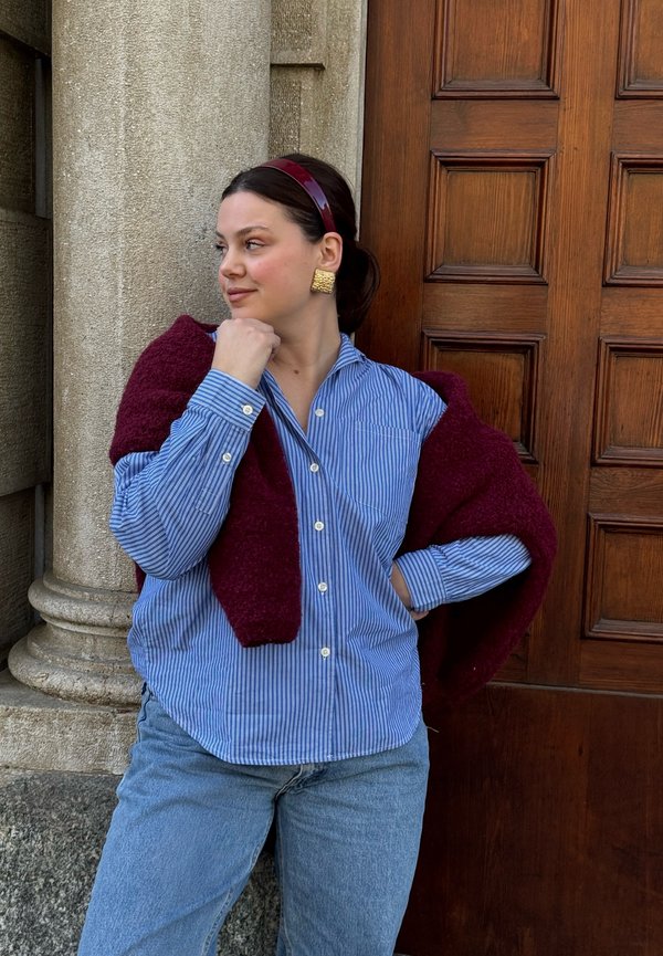 Jonge vrouw met een blauw gestreept shirt en een kastanjebruwe trui over haar schouders, staand tegen een stenen pilaar en een houten deur.