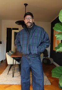 Mann iført rutete jakke, blå jeans, svart caps og briller står i en spisestue med et bord, stoler og en stor grønn plante i nærheten.