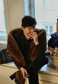 Brun imiteret pelsvikle over en sort skjorte, siddende med hånden på hagen. Højhælede sko, mørke jeans og en taske med produktæsker i nærheden.