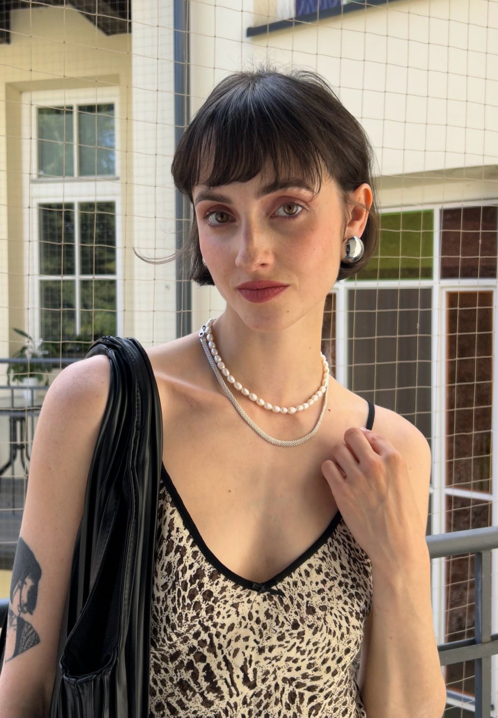 Robe à imprimé léopard avec des accents noirs, colliers en argent et perles superposés, sac noir et grandes boucles d'oreilles circulaires. Peau lisse, cheveux courts.