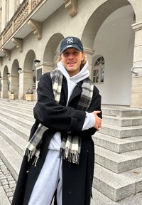 Osoba v čiernom kabáte cez šedú mikinu, s čiernou čiapkou a károvaným šálom. Na sebe má šedé tepláky, v pozadí sú kamenné schody.
