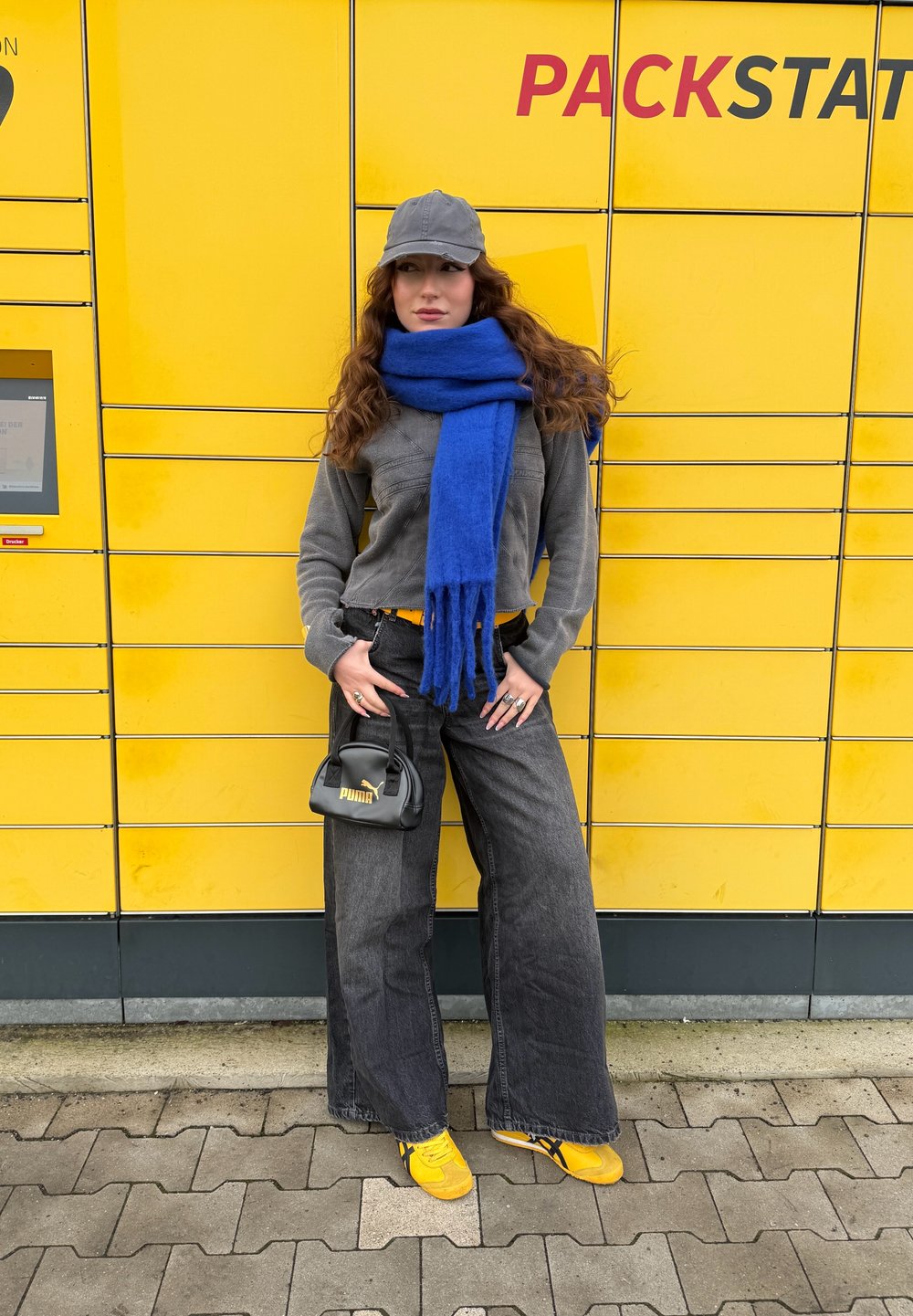 Sweater cinza de fleece, lenço azul, bolsa preta com logótipo, calças largas escuras, e sapatos amarelos em frente a um cofre de encomendas amarelo.