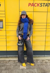 Sweater cinza de fleece, lenço azul, bolsa preta com logótipo, calças largas escuras, e sapatos amarelos em frente a um cofre de encomendas amarelo.