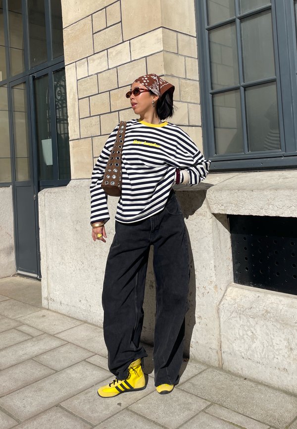 Vrouw die een gestreept shirt met lange mouwen, zwarte wijde broek, gele sneakers, een bruine bandana en een zonnebril draagt, staand tegen een stenen gebouwmuur.