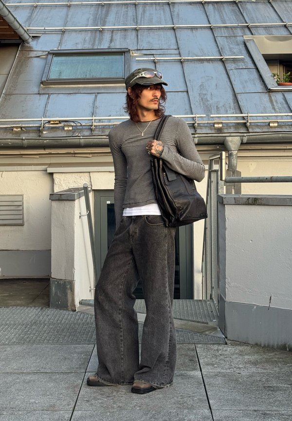 Jeune homme portant une chemise à rayures à manches longues, un jean noir large, une casquette avec des lunettes de soleil, tenant un sac en cuir noir, debout sur un toit urbain.