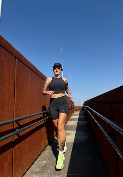 Mujer con tatuajes corriendo por un camino al aire libre con paredes de color óxido bajo un cielo azul claro, vistiendo ropa deportiva negra y zapatillas de neón.