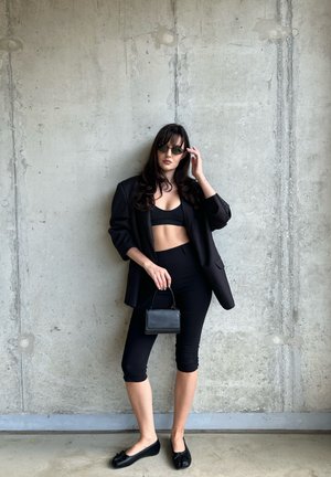 Blazer negro sobre un top corto negro y leggings, combinado con bailarinas negras. Sosteniendo un pequeño bolso negro. Fondo de concreto texturizado.