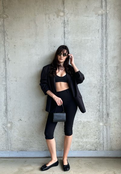 Blazer negro sobre un top corto negro y leggings, combinado con bailarinas negras. Sosteniendo un pequeño bolso negro. Fondo de concreto texturizado.
