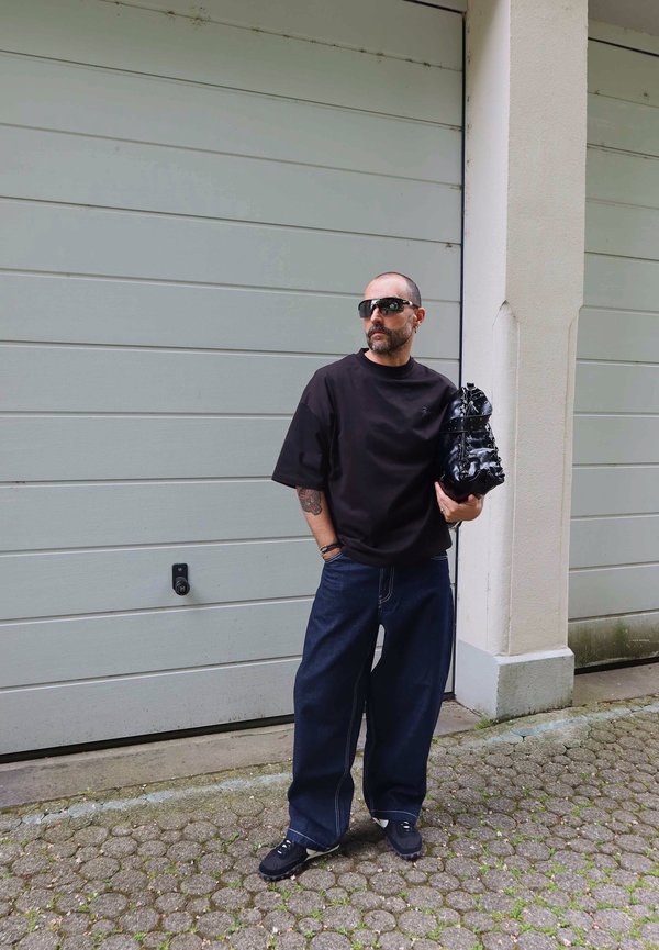 Homme portant une chemise noire oversize, un jean bleu large, des baskets noires et des lunettes de soleil, tenant un sac noir, debout sur un chemin pavé de pierres.