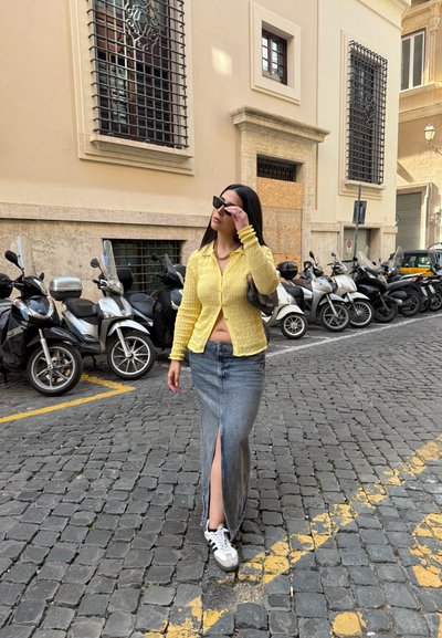 Cardigan texturizado amarillo, falda maxi de mezclilla con una abertura frontal y zapatillas blancas. Escena ambientada en una calle de adoquines con scooters estacionados.