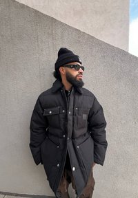 Μαύρο φουσκωτό μπουφάν με κουμπιά στερέωσης, πλαϊνές τσέπες και γιακά με γούνα. Φοριέται με μια μαύρη σκουφίτσα και γυαλιά ηλίου μπροστά σε μια υφή τοίχου.