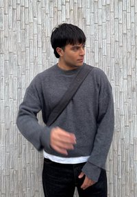 Άνδρας που φοράει γκρι πουλόβερ και μαύρη τσάντα χιαστί στέκεται μπροστά σε γκρι τοιχο με υφή, κοιτάζοντας προς τα δεξιά, με το αριστερό του χέρι σηκωμένο.