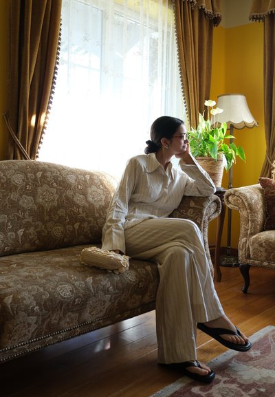 Mujer con conjunto blanco a rayas y gafas sentada en un sofá floral marrón junto a una ventana con cortinas translúcidas, sosteniendo un bolso de mano beige.