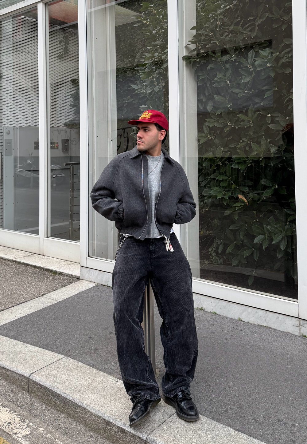 Giubbotto bomber grigio, maglione grigio chiaro, jeans larghi neri, scarpe nere, cappellino rosso. In piedi contro un vetro con vegetazione sullo sfondo.
