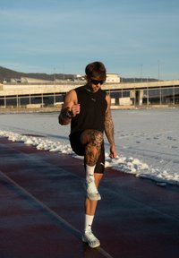 Αθλητής με τατουάζ που φοράει μαύρα αθλητικά ρούχα και γυαλιά ηλίου σηκώνει το γόνατο ενώ κάνει προθέρμανση σε χιονισμένη εξωτερική πίστα στο ηλιοβασίλεμα.