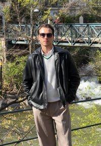 Juodas odinis švarkas ant balto tekstūruoto megztinio su žaliais akcentais, derinamas su šviesiai rudais kelnėmis. Akiniai nuo saulės ir natūralus peizažas.