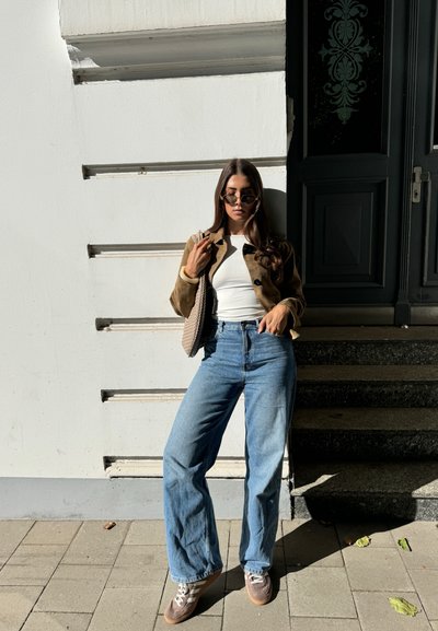 Chaqueta de ante marrón sobre una blusa blanca, jeans de mezclilla de tiro alto de color azul claro con campana, y zapatillas marrones con acentos crema. Bolso de mimbre marrón.