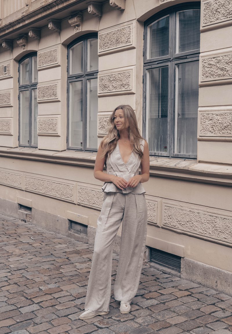 Femme aux longs cheveux portant un haut sans manches de couleur claire et un pantalon large, debout dans une rue pavée près d’un bâtiment beige avec de grandes fenêtres.