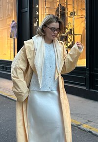 Langer, hellgelber Mantel über einer grauen Strickjacke und einem schimmernden weißen Kleid; Straßenansicht mit einem Schaufenster im Hintergrund.