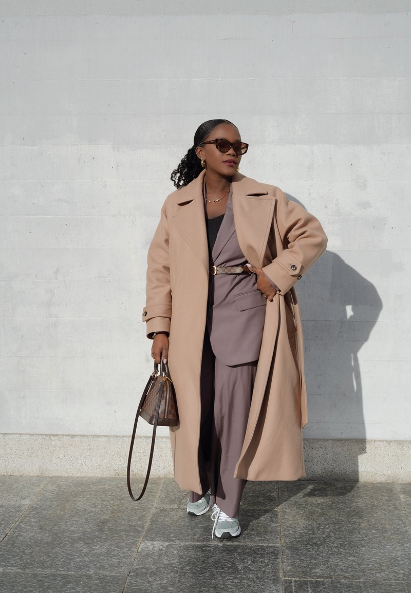 Μπεζ παλτό από μείγμα μαλλιού, φορεμένο πάνω από ένα καφέ κοστούμι. Γκρι αθλητικά παπούτσια με λευκές κορδόνες. Καφέ τσάντα χειρός με σχέδιο. Τα αξεσουάρ περιλαμβάνουν γυαλιά.