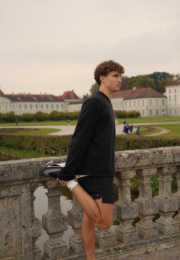 Un jeune homme vêtu de vêtements de sport noirs étire une jambe vers l’arrière près d’une balustrade en pierre, dans un jardin à la française.