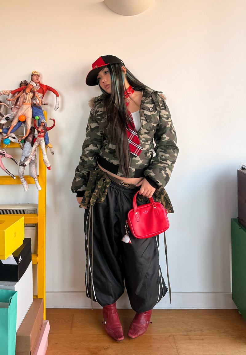 Jeune femme en veste camouflage, cravate à carreaux rouges, pantalon noir ample, bottes rouges et sac à main rouge, debout à côté d'une étagère jaune avec des poupées suspendues.
