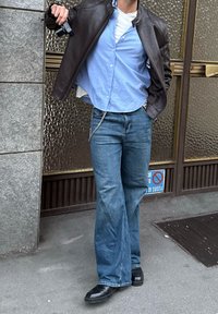 Καφέ δερμάτινο μπουφάν πάνω από ένα ανοιχτό μπλε πουκάμισο με κουμπιά, σε συνδυασμό με μπλε φαρδιά τζιν και μαύρα παπούτσια. Λεπτομέρεια αλυσίδας στα τζιν.