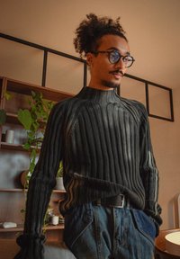 Homme aux cheveux bouclés et lunettes, portant un pull côtelé foncé et un jean, debout près d'une étagère avec des plantes et des bougies à l'intérieur.