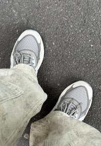 Grå sportskor med texturerad mesh och släta läderdetaljer, snörda med platta grå skosnören. Ljusfärgade byxor är synliga.