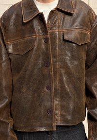 Bruine leren jas met een gestructureerde afwerking, twee borstzakken en vijf knopen in een contrasterende kleur. Verkort ontwerp met een kraag.