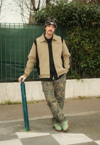 Veste en velours côtelé beige et noire, chemise noire à boutons, pantalon camouflage, chaussures vertes, debout à côté d'un poteau bleu, verdure en arrière-plan.