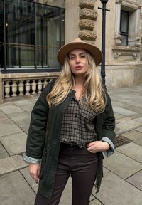 Dunkelgrüne, knöpfbare Jacke mit karierten Ärmeln über einem braunen karierten Hemd, kombiniert mit dunkelbraunen Hosen und einem beigefarbenen Filzhut.