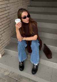 Egy személy szürke lépcsőkön ül, barna bordázott pulóvert és bőszárú kék farmert visel, fehér kávéscsészét tart a kezében, fekete mokaszinokkal és barna válltáskával.