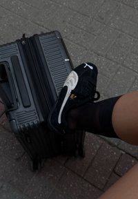 Valigia nera con ruote e superficie strutturata, accanto a una sneaker nera e argento. La sneaker presenta un logo ed è abbinata a un calzino nero.