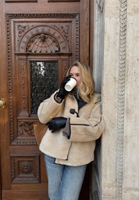 Bézs színű, fekete szegéllyel d�íszített szőrmés kabát, alatt sötét felső; fekete kesztyű. Világos koptatott farmer. Fehér csészét tart egy faragott mintás faajtó mellett.