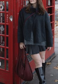 Žena u tamnom kaputu i suknji drži bordo pletenu torbu, stojeći pored crvene britanske telefonske govornice na pločniku prekrivenom opalim lišćem.