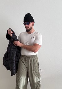 Homme portant un bonnet noir et des lunettes de soleil, un t-shirt moulant blanc Nike, et un pantalon beige, tenant une veste matelassée noire contre un mur blanc.